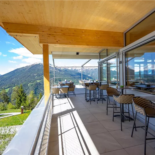 Eine moderne Terrasse mit Stühlen und Tischen bietet einen Blick auf die Berge. Im Hintergrund sind grüne Wiesen und ein klarer blauer Himmel zu sehen.