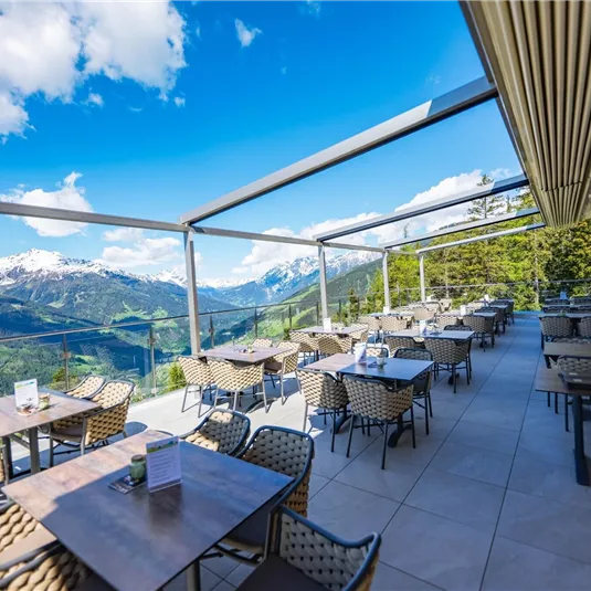 Eine moderne Terrasse mit Tischen und Stühlen, umgeben von einer atemberaubenden Berglandschaft. Der Himmel ist klar und die Aussicht bietet schöne, schneebedeckte Gipfel.