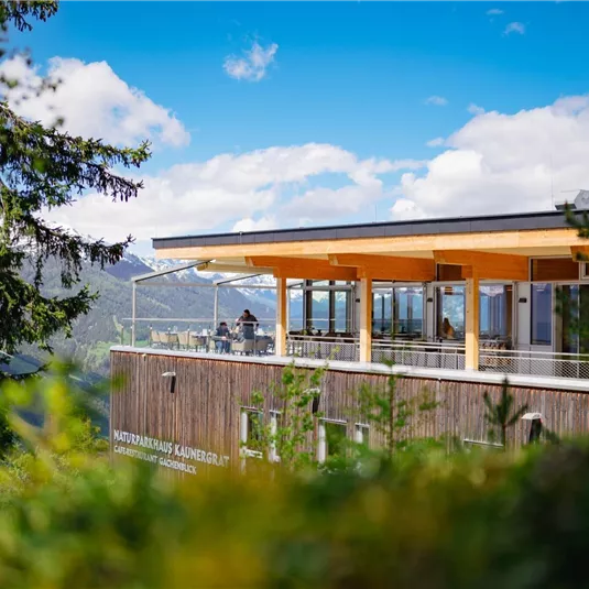Ein modernes Gebäude in den Bergen mit großen Fenstern und einer Terrasse. Der Himmel ist blau und teilweise bewölkt, umgeben von Bäumen.