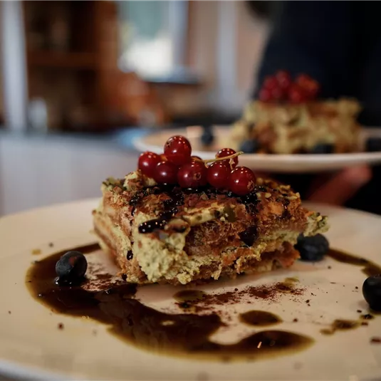 A piece of cake on a plate, garnished with red currants and blueberries. Alongside, a sweet sauce on the plate.