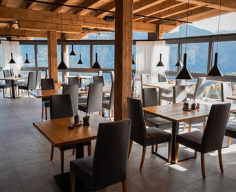 A modern restaurant with large windows and wooden elements. The tables and chairs are simple and elegant, with a view of the mountains.
