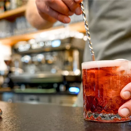 A bartender stirs a cocktail in a glass. Next to him is a carafe with another drink measure.