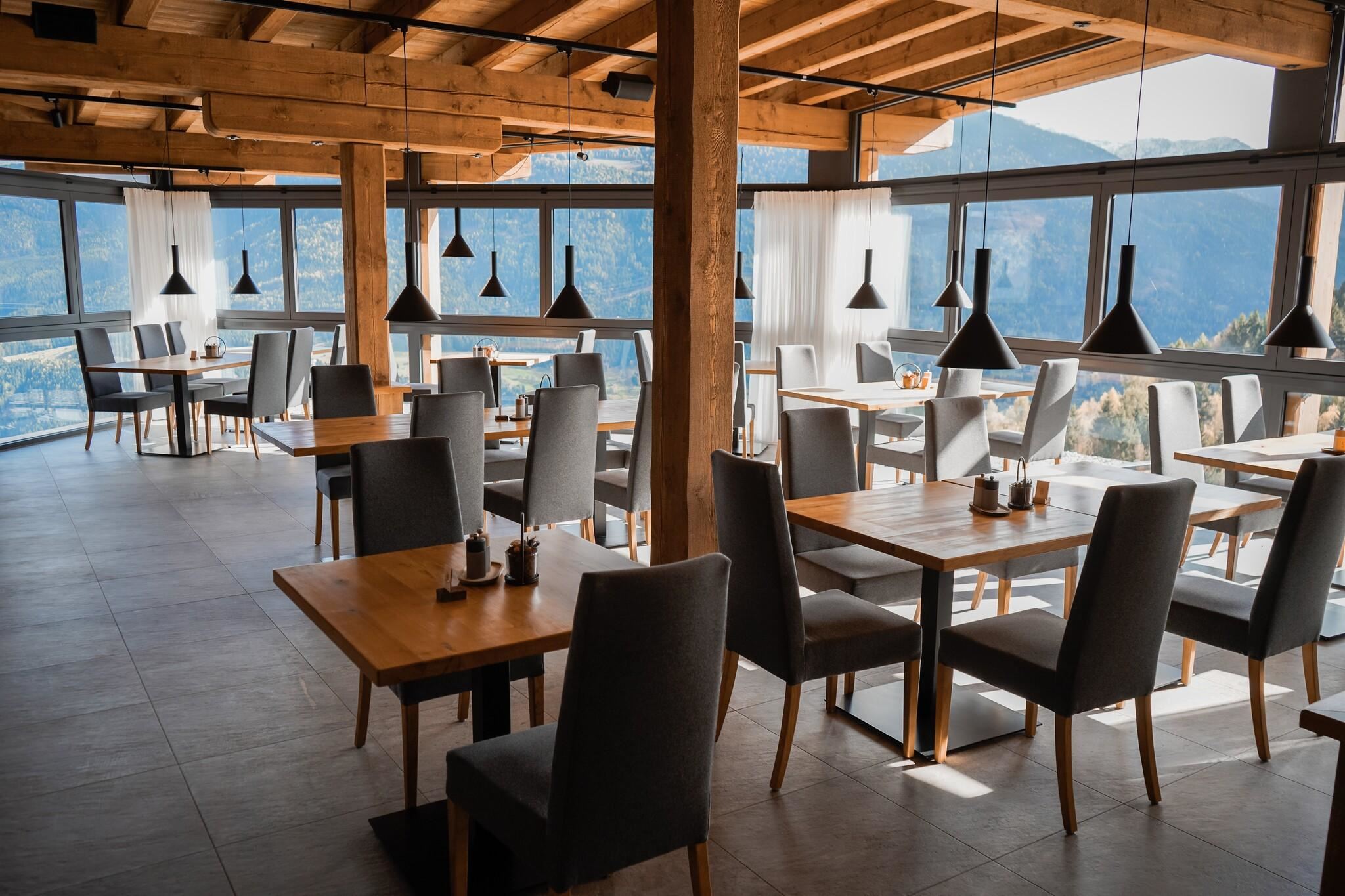 A modern restaurant with large windows and wooden elements. The tables and chairs are simple and elegant, with a view of the mountains.
