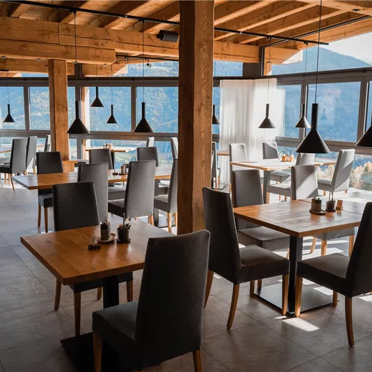 A modern restaurant with large windows and wooden elements. The tables and chairs are simple and elegant, with a view of the mountains.