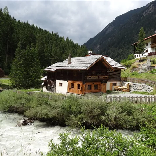 Ein rustikales Haus inmitten von Bergen und Bäumen. Ein klarer Fluss fließt neben dem Grundstück.