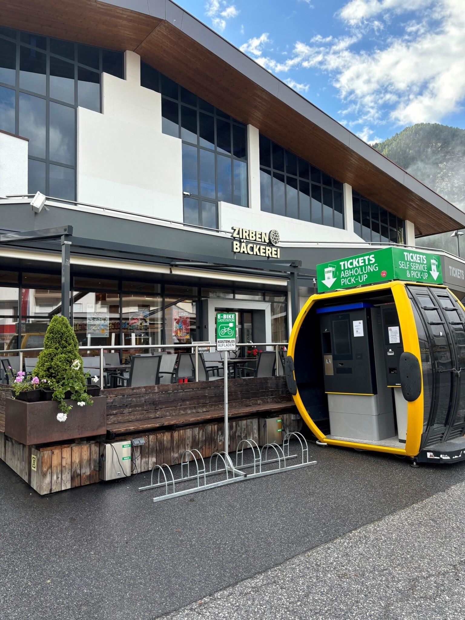 Eine moderne Bäckerei mit Außensitzplätzen und großen Fenstern. Vor dem Eingang steht ein gelbes Ticketautomat.