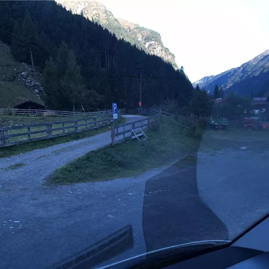 Eine ruhige Berglandschaft mit einem schmalen Weg und uhren Weidezaun. Im Hintergrund sind hohe Berge und einige Gebäude sichtbar.