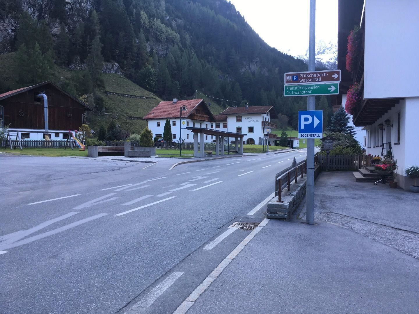 Ein ruhiger Dorfplatz mit wenigen Häusern und einer Straße. Im Hintergrund sind hohe Berge und grüne Wiesen zu sehen.