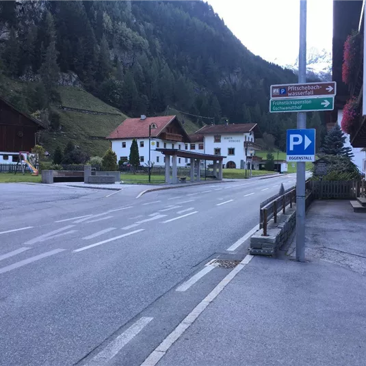 Ein ruhiger Dorfplatz mit wenigen Häusern und einer Straße. Im Hintergrund sind hohe Berge und grüne Wiesen zu sehen.