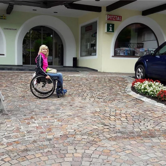 Eine Person im Rollstuhl sitzt auf einem gepflasterten Platz vor einem Gebäude. Im Hintergrund steht ein blauer Wagen und bunte Blumenbeete sind zu sehen.