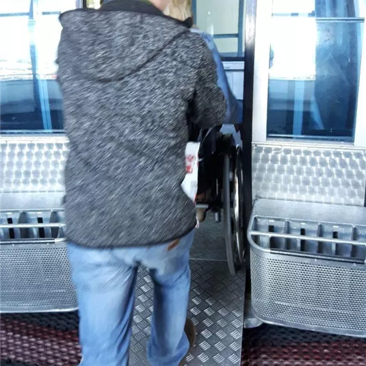 Eine Person hilft jemandem im Rollstuhl, in eine Seilbahn einzusteigen. Die Szenerie zeigt den Eingang zur Kabine mit einem rampenförmigen Zugang.