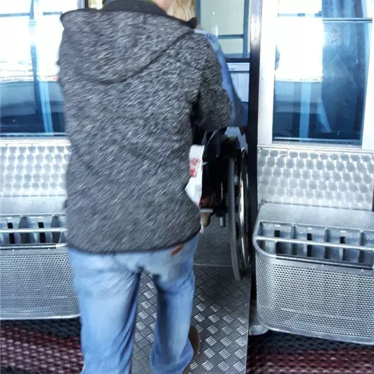 Eine Person hilft einer anderen Person im Rollstuhl, in eine Gondel einzusteigen. Die Umgebung ist modern und gut zugänglich gestaltet.
