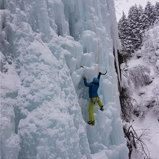 Ein Eiskletterer, der an einer glitzernden Eiswand hochklettert. Die Umgebung ist schneebedeckt und winterlich.
