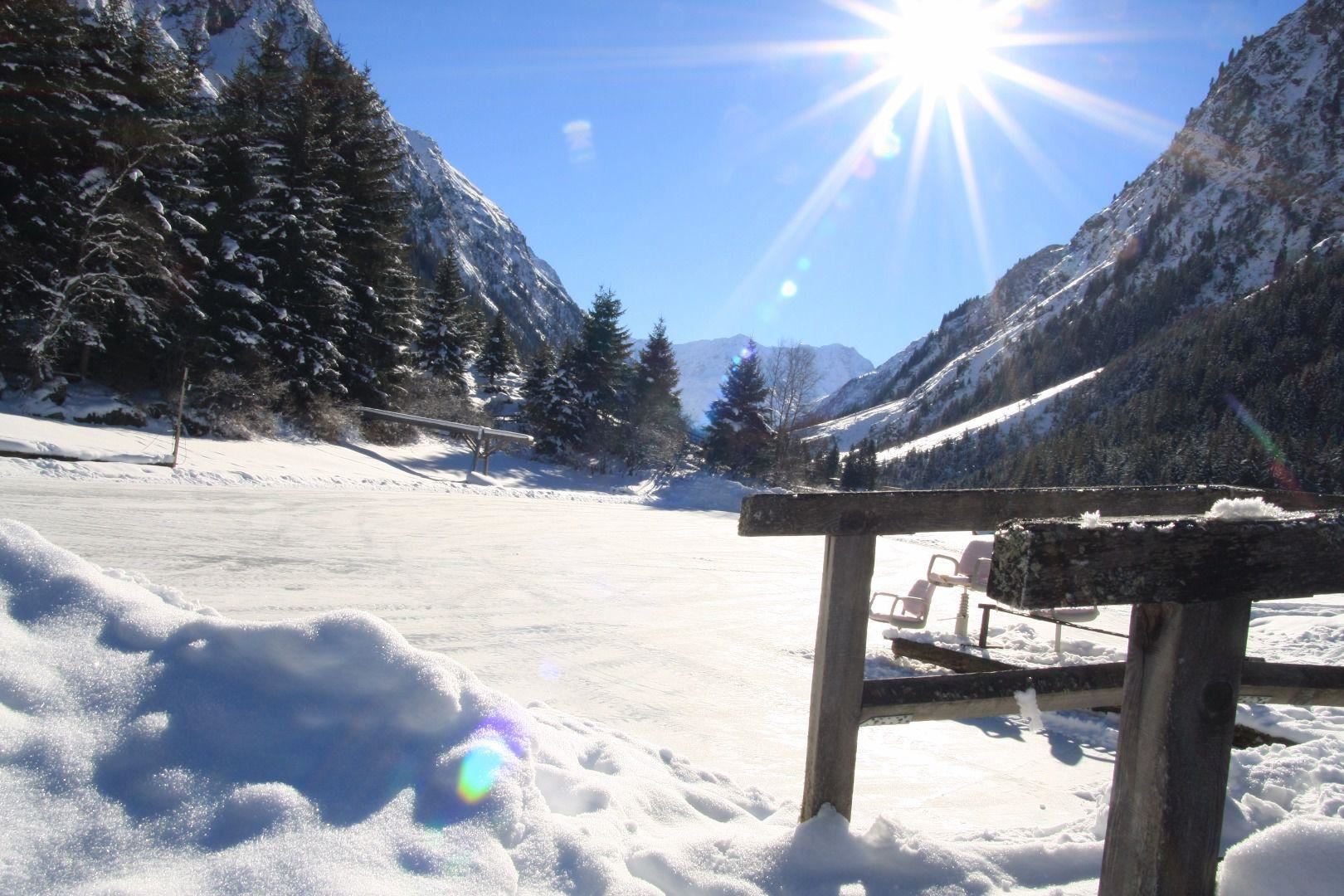 Eine verschneite Landschaft mit hohen Bergen und Nadelbäumen. Die Sonne strahlt hell über das Winterwunderland.