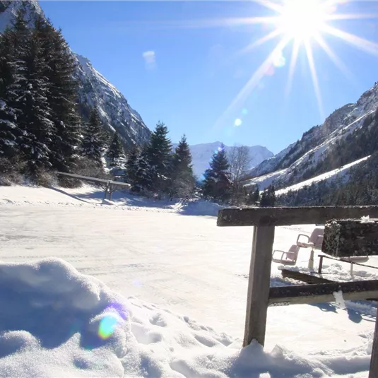 Eine verschneite Landschaft mit hohen Bergen und Nadelbäumen. Die Sonne strahlt hell über das Winterwunderland.