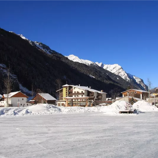 Eine schneebedeckte Landschaft mit Bergen im Hintergrund. Im Vordergrund sind Häuser zu sehen, umgeben von glattem Eis.