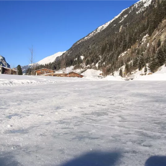 Eine verschneite Landschaft mit einem zugefrorenen See und hohen Bergen im Hintergrund. Der Himmel ist klar und blau.