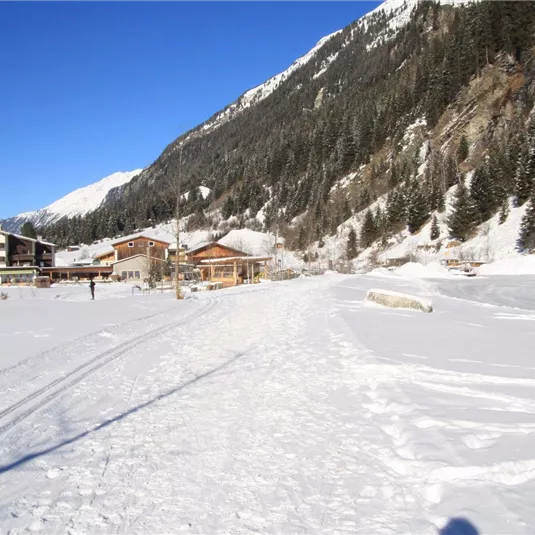 Eine winterliche Landschaft mit viel Schnee und schneebedeckten Bergen. Im Hintergrund sind einige rustikale Gebäude sichtbar.