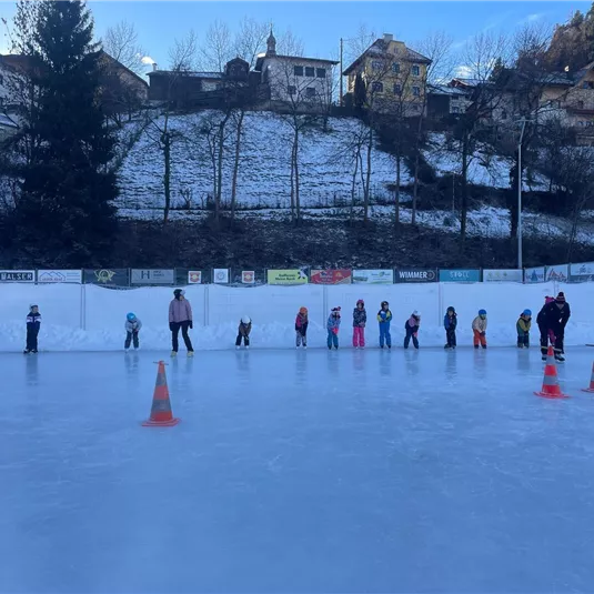 Eine Gruppe von Kindern übt das Eislaufen auf einer Eisbahn. Im Hintergrund sind schneebedeckte Hügel und Häuser zu sehen.