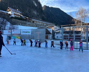 A group of children is practicing ice skating on an ice rink. In the background, some buildings and mountains can be seen.