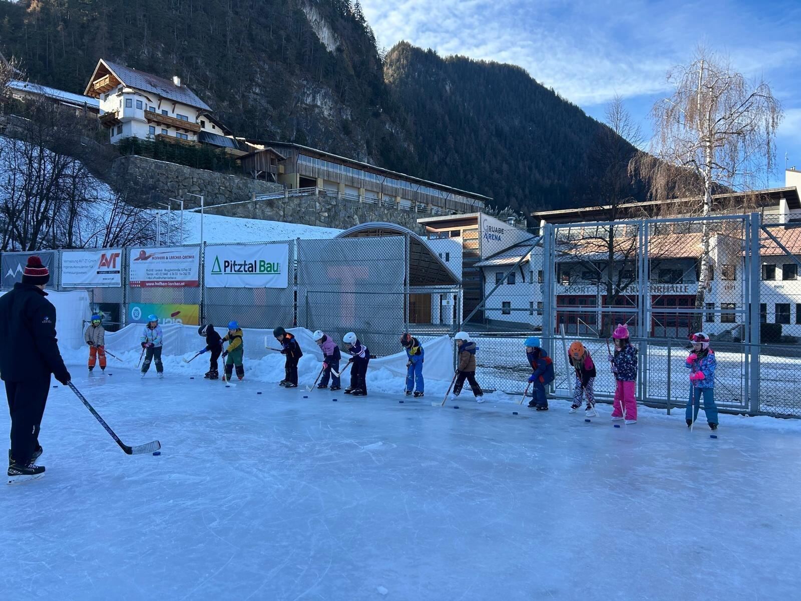 Eine Gruppe von Kindern übt das Eislaufen auf einer Eisbahn. Im Hintergrund sind einige Gebäude und Berge zu sehen.