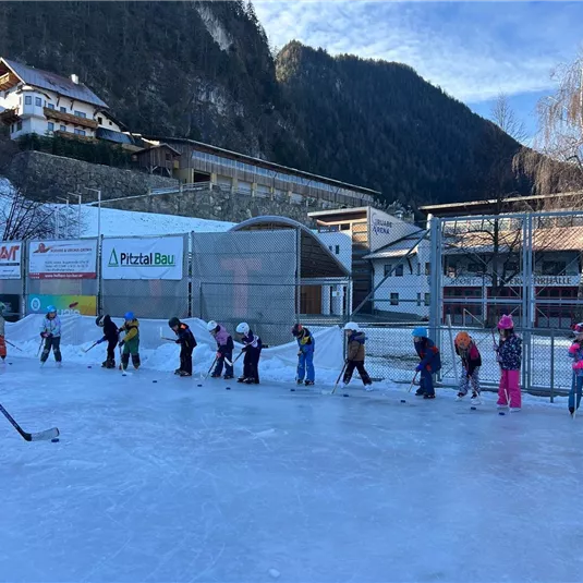 Eine Gruppe von Kindern übt das Eislaufen auf einer Eisbahn. Im Hintergrund sind einige Gebäude und Berge zu sehen.