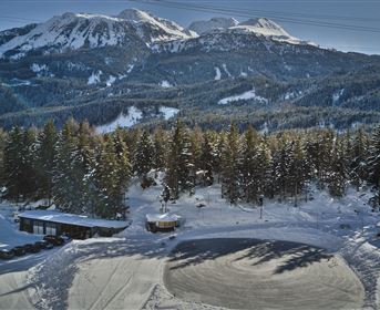 Eine winterliche Landschaft mit schneebedeckten Bergen und dichten Nadelbäumen. Im Vordergrund sind einige Hütten und ein abgerundeter Platz im Schnee sichtbar.