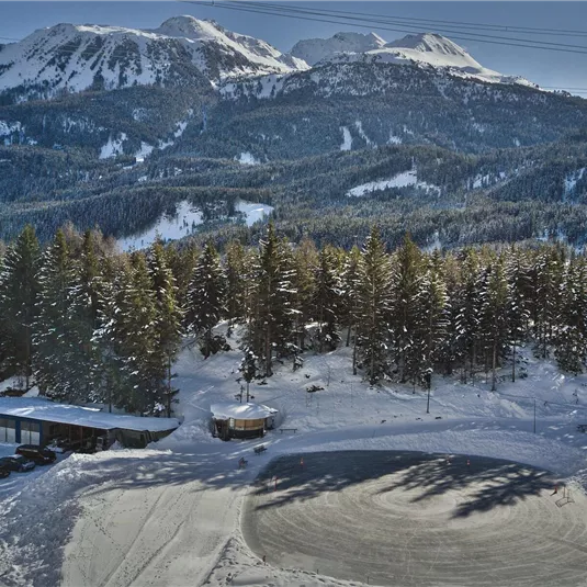Eine schneebedeckte Landschaft mit majestätischen Bergen im Hintergrund. Im Vordergrund sind kleine Hütten und eine Piste zu sehen.