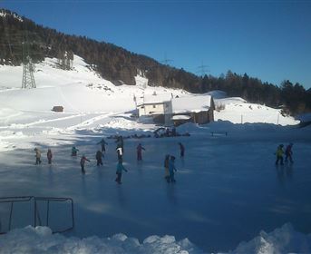 Ein gefrorener See mit vielen Eisläufern in winterlicher Landschaft. Im Hintergrund sind schneebedeckte Berge und ein blauer Himmel zu sehen.