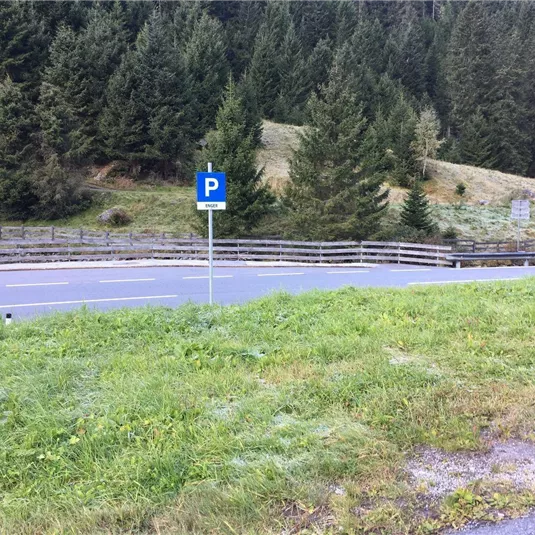 Eine Parkfläche neben einer Straße, umgeben von Bäumen. Im Hintergrund ist eine grüne Wiese sichtbar.