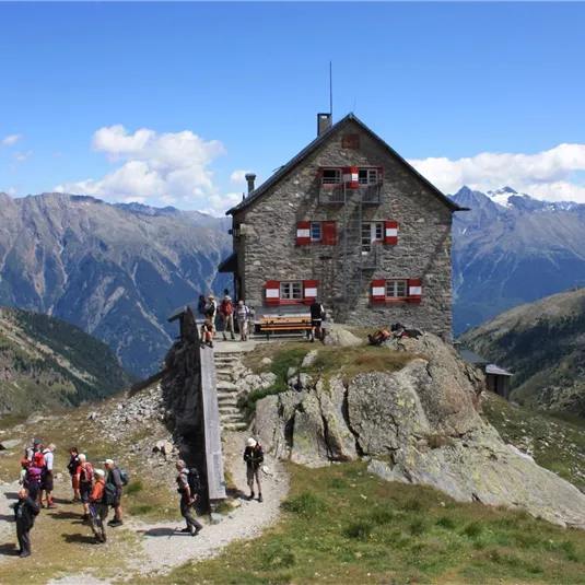 A stone mountain house with red windows, surrounded by a spectacular mountain landscape. Hikers gather in front of the house and enjoy the view.