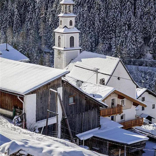 Ein malerisches Bergdorf im Winter mit schneebedeckten Häusern. Im Hintergrund ist eine Kirchturm mit einer schönen Baumkullisse sichtbar.