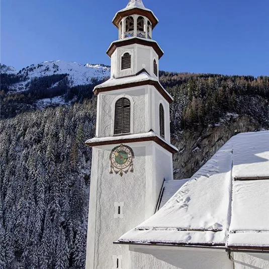 Ein kirchlicher Turm steht in der Winterlandschaft, umgeben von schneebedeckten Bäumen. Der Himmel ist klar und blau, was der Szene eine friedliche Atmosphäre verleiht.