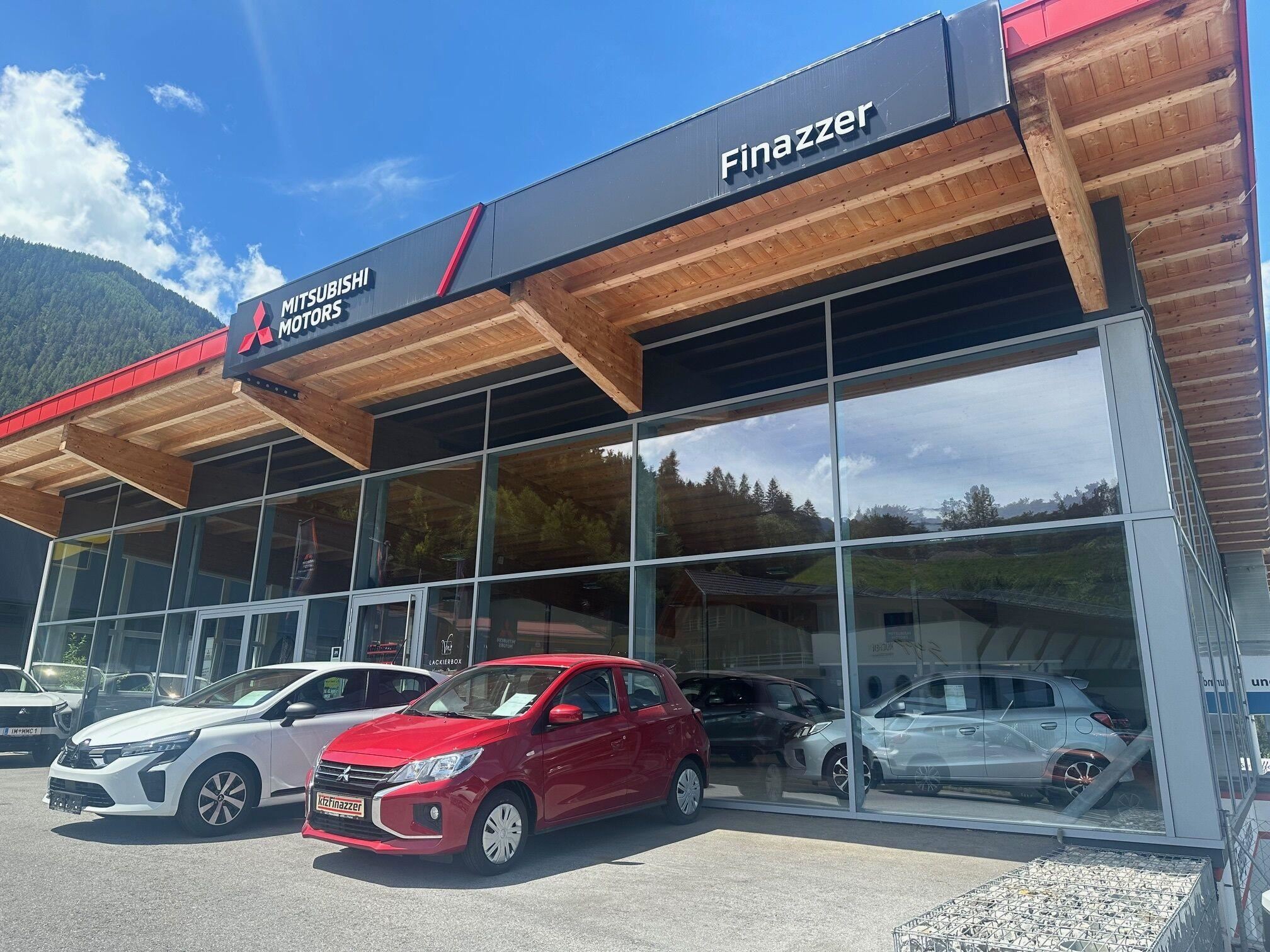 Eine Mitsubishi-Autoverkaufsstelle mit mehreren Fahrzeugen vor einem modernen Gebäude. Der Himmel ist blau und die Umgebung ist von Bergen umgeben.