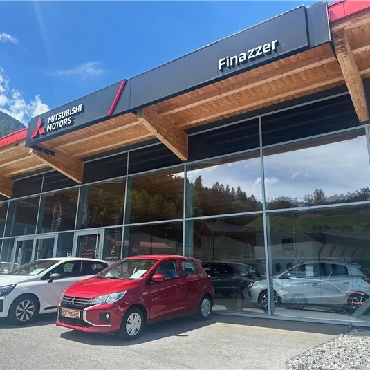 Eine Mitsubishi-Autoverkaufsstelle mit mehreren Fahrzeugen vor einem modernen Gebäude. Der Himmel ist blau und die Umgebung ist von Bergen umgeben.