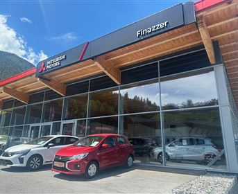 Ein Autohaus mit modernen Fahrzeugen vor großen Glasfenstern. Die Umgebung zeigt Berge und blauen Himmel.