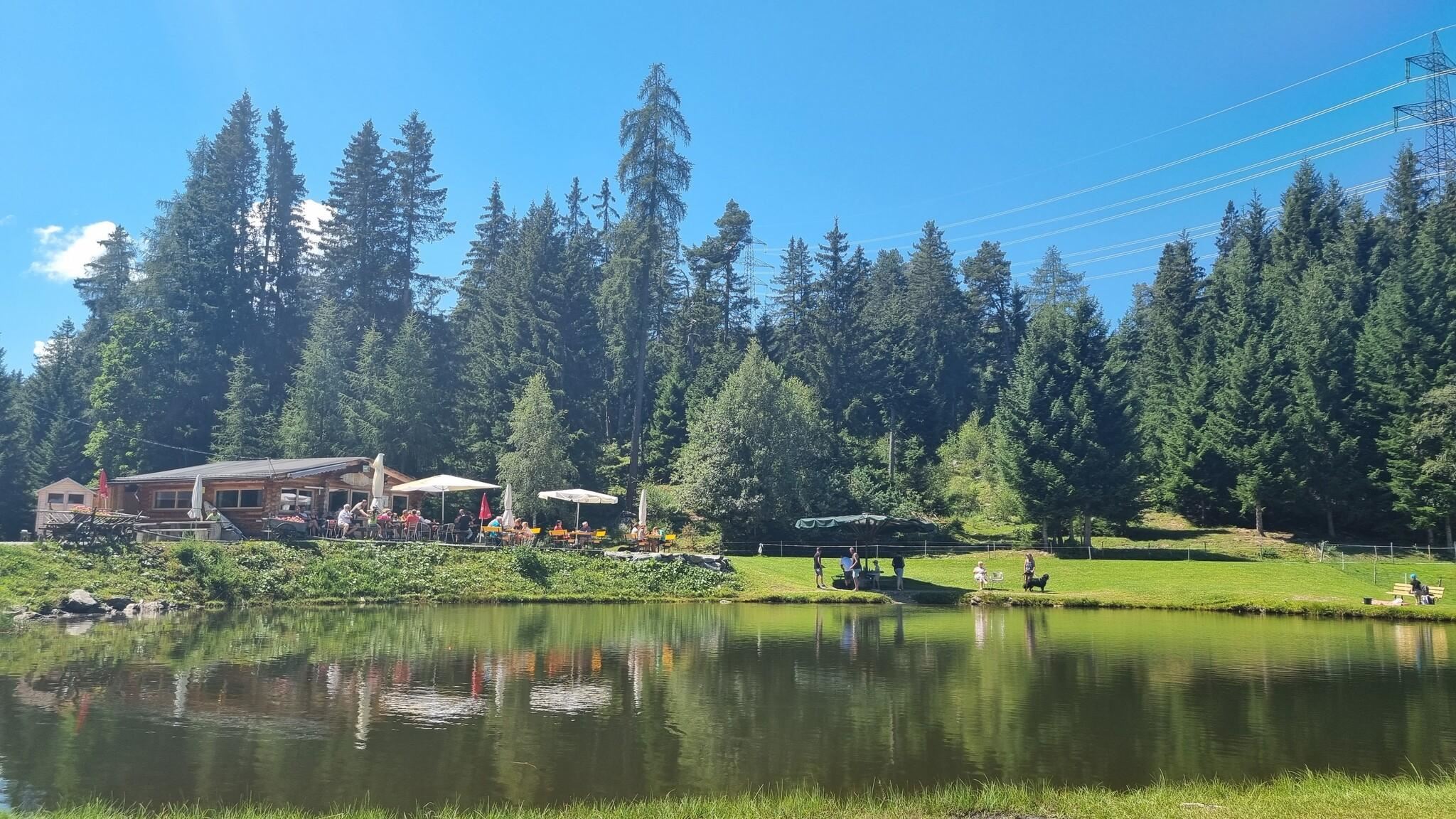 A picturesque place by the lake surrounded by tall trees. In the background, an inn and outdoor seating can be seen.