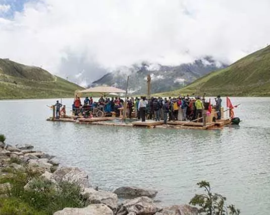 Eine große Gruppe von Menschen steht auf einer Holzplattform im Wasser. Im Hintergrund sind grüne Hügel und bewölkter Himmel zu sehen.