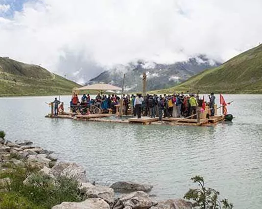 Eine große Gruppe von Menschen steht auf einer Holzflotte auf einem See. Im Hintergrund sind grüne Berge und bewölkter Himmel zu sehen.