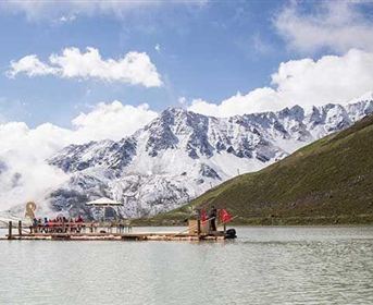 A floating platform on a clear lake, surrounded by snow-covered mountains. The sky is overcast, and the landscape is wild and picturesque.
