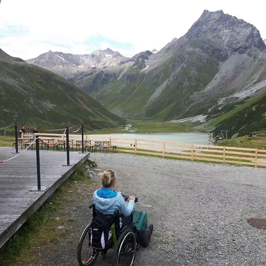 Eine Person im Rollstuhl fährt auf einem Schotterweg durch eine malerische Berglandschaft. Im Hintergrund sind hohe Berge und ein ruhiger See zu sehen.
