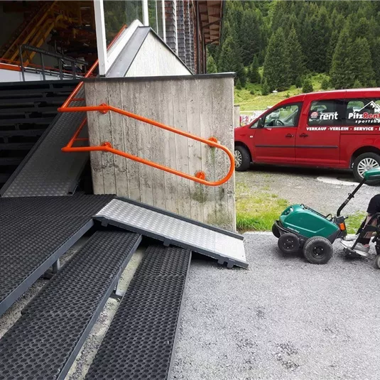 Eine Rampe neben einer Treppe und ein rotes Fahrzeug im Hintergrund. Im Vordergrund ist ein Rollstuhl zu sehen.