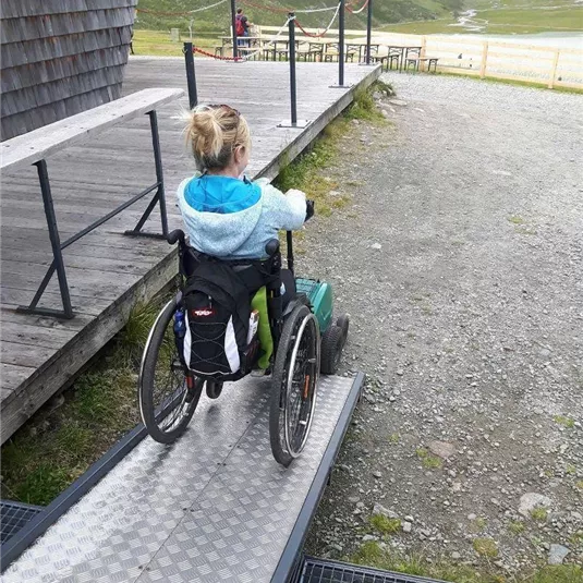 Ein Kind im Rollstuhl fährt eine Rampe hinauf. Die Umgebung ist von Bergen und Grünflächen umgeben.
