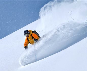 Ein Skifahrer in leuchtend orangefarbener Kleidung fährt durch frischen, tiefen Schnee. Der klare blaue Himmel sorgt für eine perfekte Winteratmosphäre.