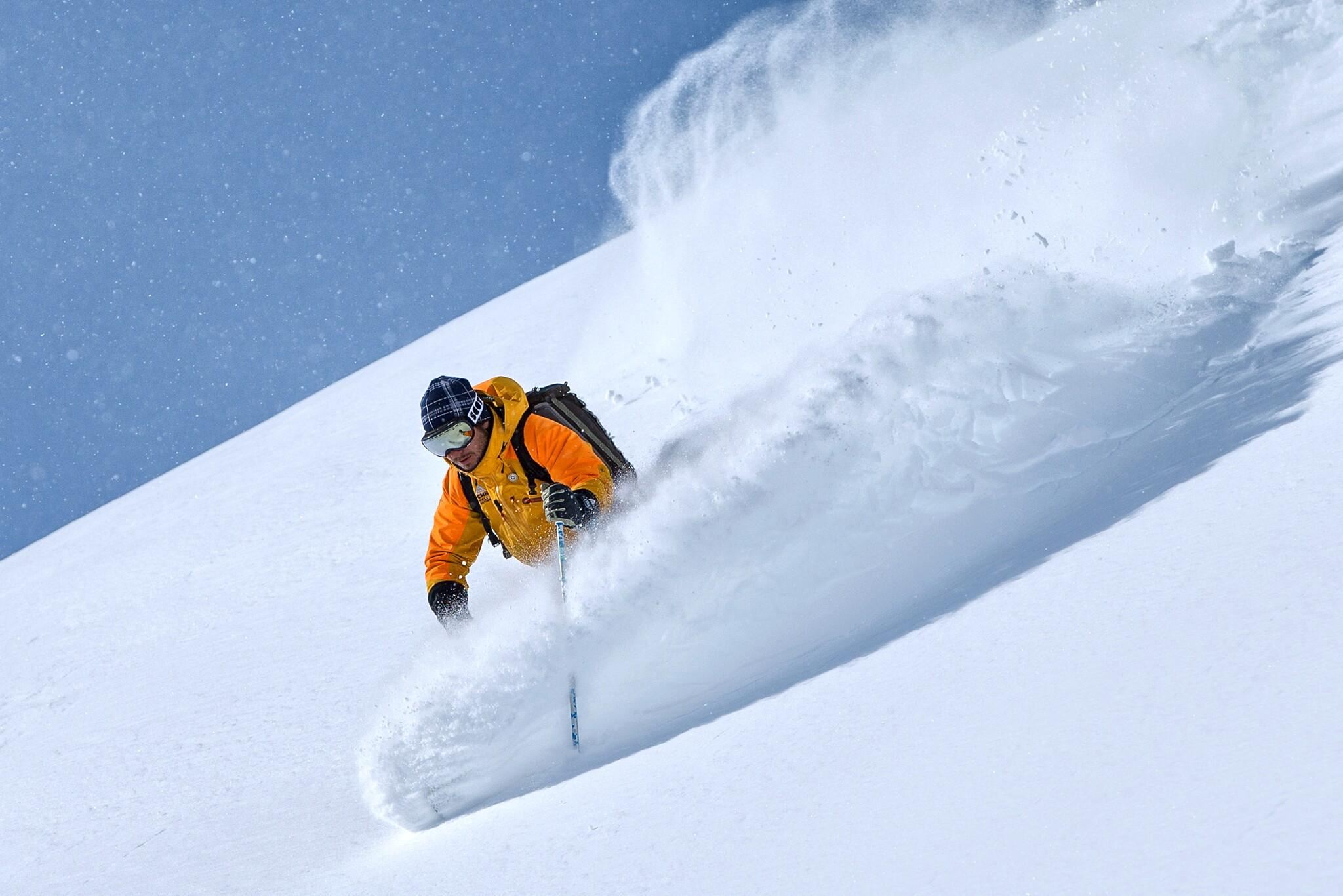 Ein Skifahrer in leuchtend orangefarbener Kleidung fährt durch frischen, tiefen Schnee. Der klare blaue Himmel sorgt für eine perfekte Winteratmosphäre.