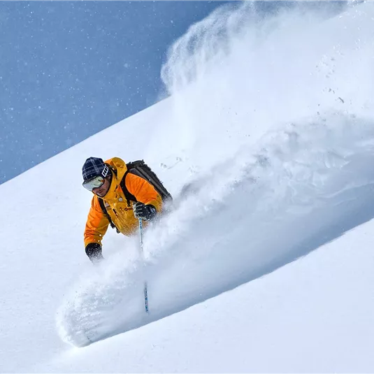 Ein Skifahrer in leuchtend orangefarbener Kleidung fährt durch frischen, tiefen Schnee. Der klare blaue Himmel sorgt für eine perfekte Winteratmosphäre.