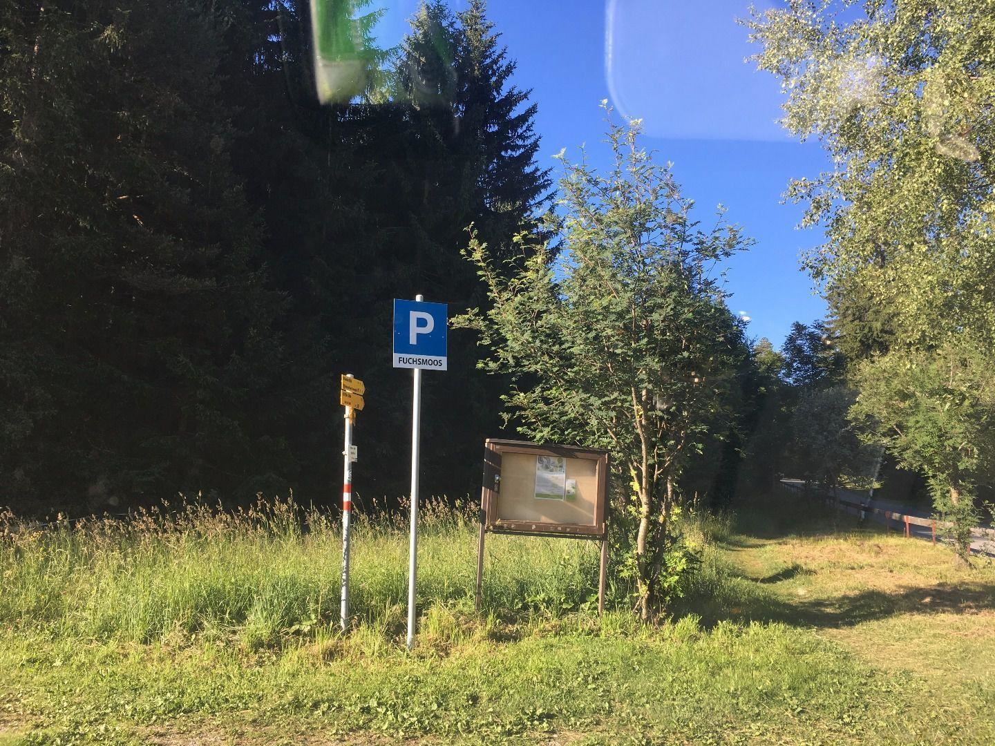 Ein Parkplatzschild steht neben einer Wiese mit Bäumen im Hintergrund. Es ist ein klarer, blauer Himmel zu sehen.