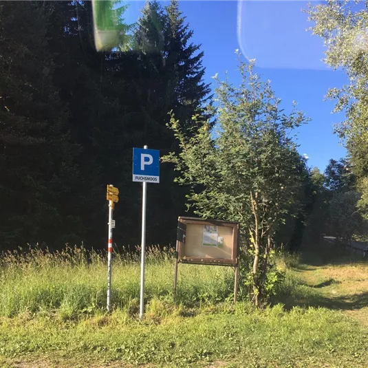 Ein Parkplatzschild steht neben einer Wiese mit Bäumen im Hintergrund. Es ist ein klarer, blauer Himmel zu sehen.