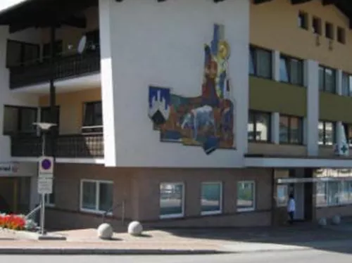 Ein modernes Gebäude mit einem großen Wandbild und Balkonen. Vor dem Gebäude sind Blumenbeete und eine Straßenansicht sichtbar.