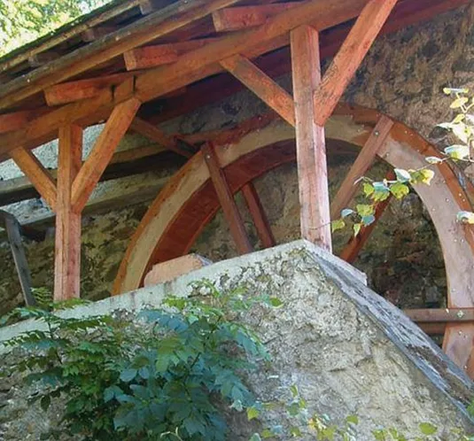 Ein Wasserrad aus Holz, das unter einem überdachten Bau steht. Umgeben von Pflanzen und einem klaren Himmel.